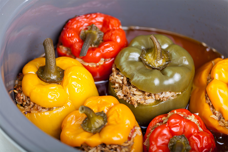 Drei gelbe, zwei rote und eine grüne Paprika gefüllt mit Reis und Faschiertem in einer ovalen grauen Auflaufform. Die Paprika sind weich vom Backen und liegen in Tomatensauce.