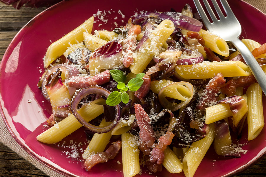 Dunkelrotes tiefes Teller darauf Penne mit Raddichio, garniert mir Parmesan und Basilikum.