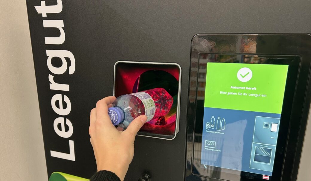 Eine Flasche wird in einen schwarzen Pfandautomaten eingeworfen. Am Bildschirm ist zu erkennen, dass der Automat bereit ist und das Leergut eingelegt werden kann.